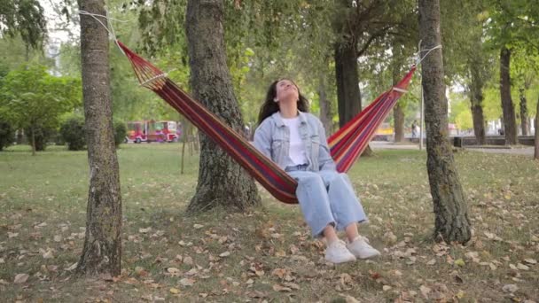 若い幸せな魅力的な女性は 公園のファブリックハンモックチェアに座ってリラックスを楽しんでいます 自然の中で平和な休息 — ストック動画