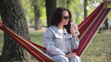 Genç bir kız su içiyor, bir sonbahar parkında hamak kullanıyor..