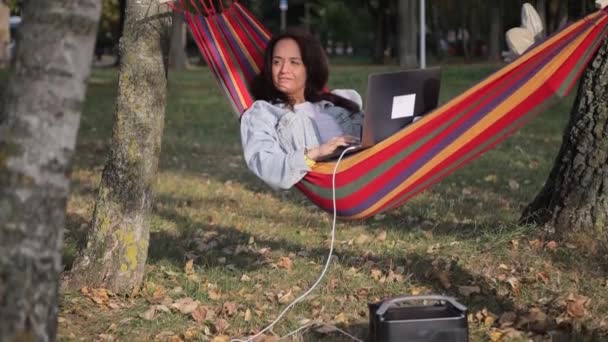 这个女孩在一台笔记本电脑上工作 这台笔记本电脑是从一个入口充电站充电的 在公园的吊床上休息 — 图库视频影像