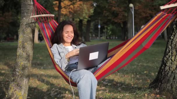 公園のハンモックでノートパソコンを使って横たわっている女性フリーランサー 新鮮な空気の中で働く — ストック動画