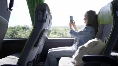 Sarışın bir kadın turist otobüsünde akıllı bir telefonla çekim yapıyor, oturuyor ve pencereden dışarı bakıyor..