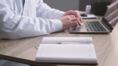Hasta bilgilerini dolduran bir kalem tutan erkek doktor ve doktor odasındaki ofis masasında dizüstü bilgisayarda çalışmak, teletıp konsepti.