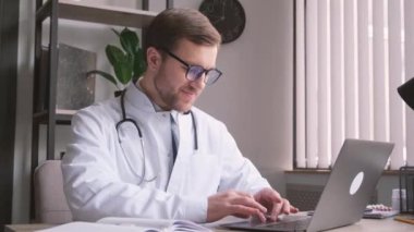 Modern bir klinikte gözlük takan ve beyaz önlüklü bir erkek doktor dizüstü bilgisayar kullanıyor, kişisel sağlık verilerini kaydediyor, uzaktan danışmanlık yapıyor. Teletıp sağlık hizmeti kavramı.