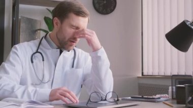 Yakışıklı genç doktor kliniğinde tek başına oturuyor ve dizüstü bilgisayarını kullanırken stresli hissediyor. Hastane ya da klinikte tıbbi stres, endişe ve yangınlar.