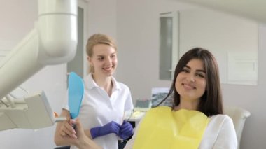 Genç mutlu kadın müşteri, dişçi muayenehanesinde dişlek bir gülümsemeyle aynaya bakıyor. Dişçi muayenehanesinde aynada dişlerini kontrol eden güzel genç bir kadın, kusursuz beyaz dişlerini gösteriyor..