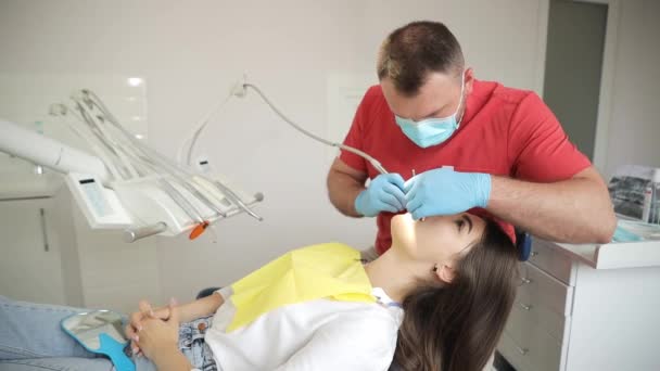 牙医非常仔细地检查和修理他的年轻病人的牙齿 戴着防护面具和手套的牙医治疗一位年轻妇女的牙齿 — 图库视频影像