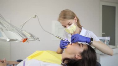 Genç bir kadın dişçide tıbbi bir sandalyede oturuyor, dişlerini bir diş kliniğinde tedavi ediyor. Kadın diş hekimi ve dişçi sandalyesinde oturan kadın diş hekimi ağız boşluğu tedavisi uyguluyor.