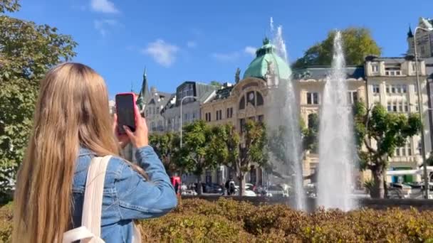 バックパックでスタイリッシュな女性旅行者は ヨーロッパの旧市街の噴水の写真を撮ります — ストック動画