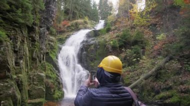 Koruyucu ekipmanlı bir turist akıllı telefondan şelalenin doğasını fotoğraflıyor. Yavaş çekim.