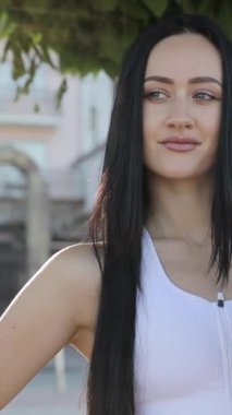 Spor kıyafetli güzel bir kız, elinde kauçuk yoga minderi, parkta duruyor. Sportif kadın yoga minderini tutuyor ve parkta antrenmana gidiyor. Dikey video