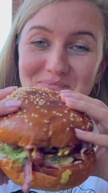 Genç bir kadın fast food restoranında et burger yiyor. Yemek yiyen ve kameraya bakan bir kadının portresi. Dikey video