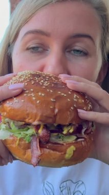 Aç bir kadın büyük bir burger yer. Sokak yemeği. Abur cubur, sosisli sandviç, hamburger, hamburger. Dikey video.