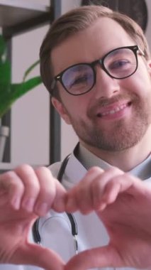 Beyaz tıbbi üniformalı gülümseyen genç pratisyen doktorun portresi. Kalp sevgisi el hareketi gösteriyor. Genç ve mutlu erkek doktor destek veriyor. İnsanları tedavi etme kavramı. Dikey video