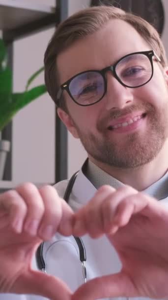 心臓愛の手のジェスチャーを示す白い医療ユニフォームで若い一般開業医を笑顔にする肖像画 サポートを示す幸せな若い男性医師 人々を扱うという概念 バーティカルビデオ — ストック動画