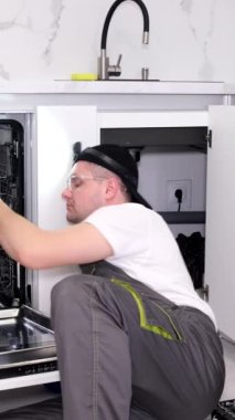Tulumlu bir tamirci onu kurduktan sonra mutfaktaki bulaşık makinesini kontrol eder. Ev aletlerinin tamiri. Dikey video.
