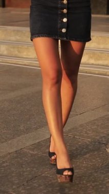 Kısa etekli ince bacaklı genç bir kız şehirde yürüyor. Dikey video. Yüksek topuklu güzel ince kadın bacakları..