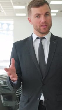 Bir erkek satıcı araba galerisinde yeni bir arabadan bahsediyor. Otomotiv işi. Dikey video.