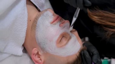 Bir güzellik uzmanı bir erkeğin yüzüne nemlendirici bir maske takar. Kozmetik kliniğindeki adam, yüz yüze..