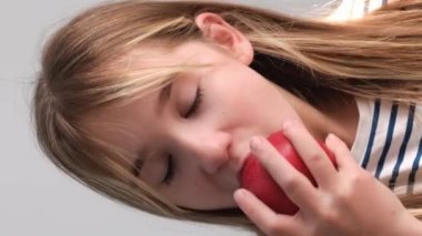 Kırmızı bir elmayı ısıran küçük tatlı bir kızın yakın çekimi. Elmayı ısıran aç güzel kız. Dikey video. İnsan duyguları, yüzler.