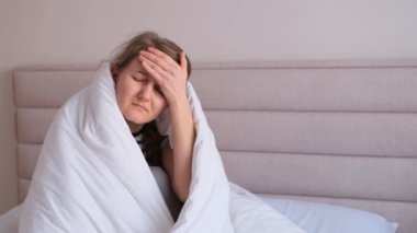 Kadın kendini iyi hissetmiyor, beyaz battaniyelerin altında yatıyor ve kaşlarını çatıyor, kafasına dokunuyor, baş ağrısı, migren ağrısı..