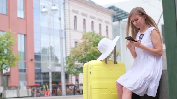 一位年轻漂亮的旅游姑娘正在用手机寻找关于公共汽车交通的信息 坐在公共汽车或电车车站的金发女旅行者 — 图库视频影像
