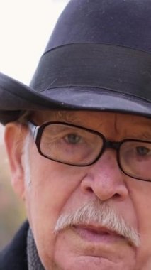 Sonbahar parkında dikilirken gözlüğünü ayarlayan şapkalı bir büyükbabanın portresi. Dikey video.