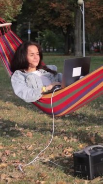 Genç bir kadın programcı bir ağacın altında dizüstü bilgisayar üzerinde çalışıyor ve onu taşınabilir şarj istasyonundan şarj ediyor. Dikey video