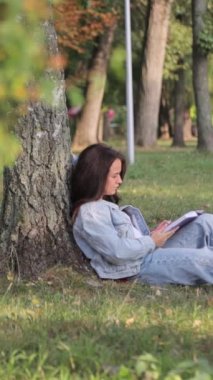 Genç bir öğrenci kız elinde bilgisayarla parkta çimlerin üzerinde oturuyor. Üniversitede okuyorum. Eğitim ve uzaktan çalışma konsepti. Dikey video