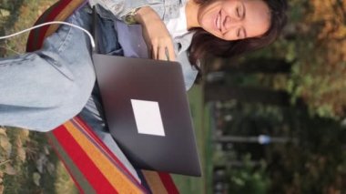 Parktaki bir hamakta dizüstü bilgisayar kullanarak yatan serbest çalışan bir kadın. Temiz havada çalış. Dikey video.