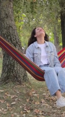 Parktaki kumaş hamak sandalyesinde oturup rahatlamanın keyfini çıkaran genç, mutlu ve çekici bir kadın. Doğada huzur içinde yatsın. Dikey video