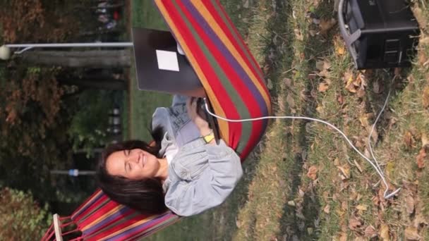 一个在夏日花园的吊床上工作的年轻女人正在一台笔记本电脑上工作 给手提式电站的笔记本电脑充电 垂直录像 — 图库视频影像