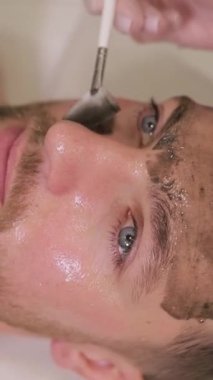 Tıraşsız bir adam kaplıca salonunda kozmetik maske takıyor. Kozmetik uzmanı spa salonunda erkeklerin yüzüne maske takıyor. Dikey video