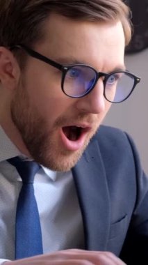 Mutlu genç iş adamı, el hareketleriyle başarıyı kutluyor, dizüstü bilgisayara bakıyor, masada oturuyor. İş adamı başarısıyla seviniyor. Dikey video.