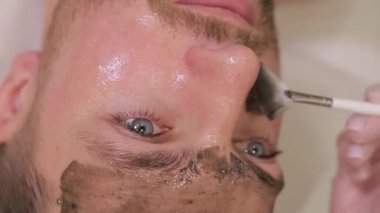 Tıraşsız bir adam kaplıca salonunda kozmetik maske takıyor. Kozmetik uzmanı spa salonunda erkeklerin yüzüne maske takıyor. Dikey video