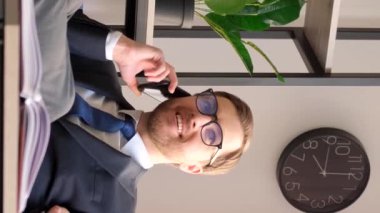 Takım elbiseli bir ofis çalışanı cep telefonuyla konuşuyor ve başarılarından bahsediyor, ofiste ayakları masanın üzerinde oturuyor. Dikey video