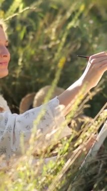 Bir sanatçı, güneşli bir yaz gününde çayırda resim çizer. Sanatçı kız doğayı çizer. Dikey video.