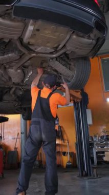 Bir araba tamircisi altında dururken bir arabanın şasesini inceler. Dikey video.