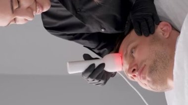 Genç bir adam güzellik kliniğinde radyo frekansı kaldırma işleminden hoşlanıyor. Spa kliniğinde yüz tedavisini yeniliyorum. Dikey video.
