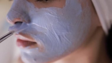 Yüzüne maske takılmış güzel bir genç kadın masaj masasında yatıyor. Makyajcı kadın derisine yüz maskesi takıyor. Kapatın. Dikey video.