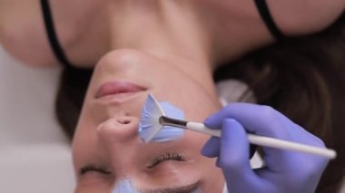 Genç kadınların yüzüne mavi kozmetoloji maskesi takan kadın kozmetikçi. Güzellik kliniğinde yüz estetiği. Dikey video.