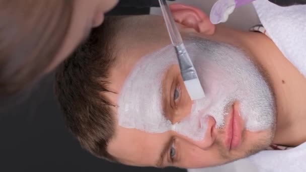 若い男性は スパサロンで美容師から栄養価の高いフェイスマスクを受け取ります スパトリートメントとスキンケア バーティカルビデオ — ストック動画