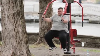 Genç, kilolu bir adam, yeşil bir alandaki açık hava egzersiz makinesinde egzersiz yapıyor. Şişman bir adam aşırı kilolarla mücadele eder. Eller için alıştırmalar.