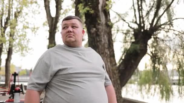一个超重的年轻人站在公园的树后的画像 一个穿着灰色T恤的胖子在室外锻炼机附近的绿地里 — 图库视频影像