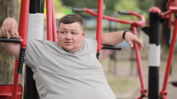 疲れた若い脂肪の男は 屋外の運動機械の上に座っている灰色のTシャツで 過剰な体重で戦っている 体重減少について — ストック動画