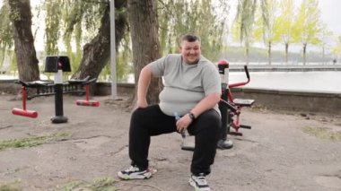 Tatmin olmuş şişman genç adam parkta egzersiz makinesine oturur. Açık hava egzersiz makinelerinin yanındaki yeşil bölgede gri tişörtlü şişman bir adam. Eğitimden sonra yorgunluk