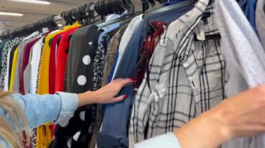 Uzun saçlı genç bir kadın bir giyim mağazasından kullanılmış giysiler alır. Bir kadın kullanılmış kıyafetleri dikkatle inceler.