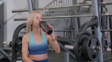 Spor kıyafetli sağlıklı bir Amerikalı kadın spor salonunda çalıştıktan sonra bir şişe protein içeceği içer..