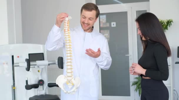 医学オフィスの解剖学的脊椎を指す医師 若い女性の患者が脊椎の脊椎について話している医師に聞きます — ストック動画