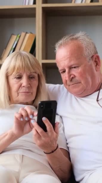 高齢のカップルは喜んでソファーに座っている間笑顔を共有し 彼らはお互いに優しく触れ ジェスチャーを交換するように携帯電話を見ます — ストック動画
