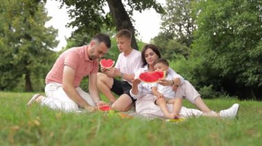 Mutlu bir aile, bir ağacın altında çimenlerde oturur, karpuzu paylaşır ve tadını çıkarır, gülümser ve doğadaki eğlence aktivitelerini hoş görür..